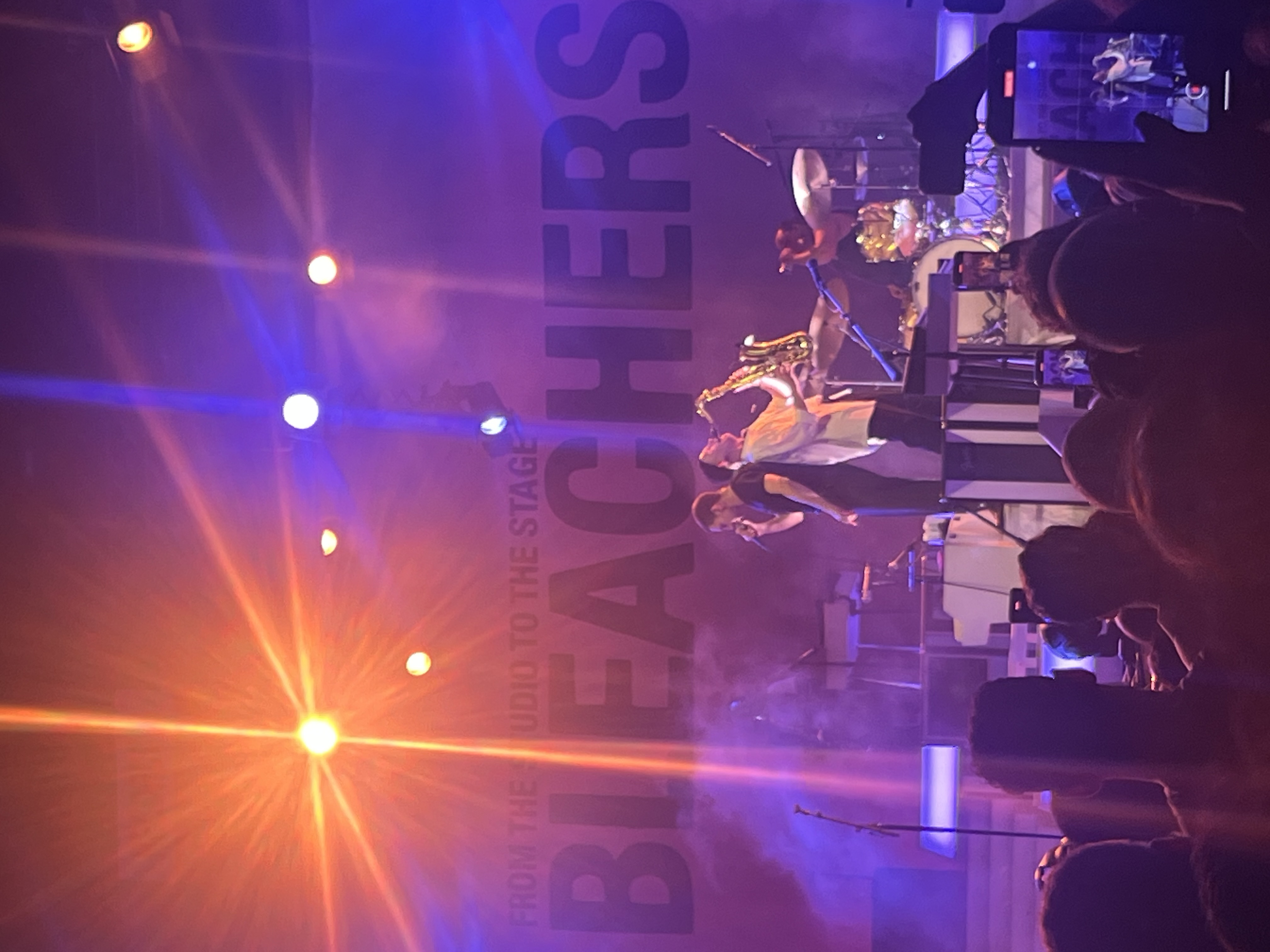 Bleachers performing on stage in New Haven, lit in orange and blue with a saxophone player center stage.